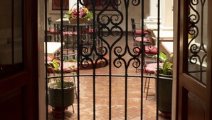 An open wrought iron gate leading to a courtyard with a tiled floor, potted plants, and a table with chairs.