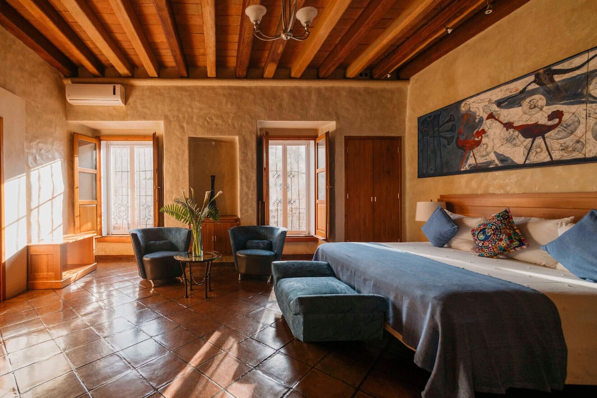 Habitación de hotel con cama, dos sillones, una mesita con un jarrón y un cuadro en la pared.