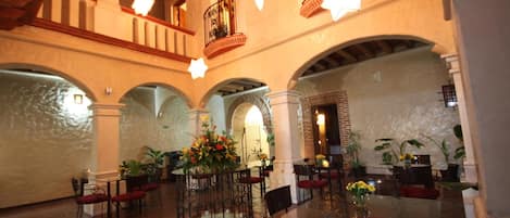 A spacious lobby with a central round table surrounded by chairs, a decorative fountain, and a balcony with a railing.