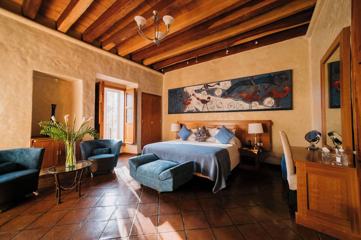 Un dormitorio amplio con una cama grande, una zona de estar con una silla y una mesa, y un cuadro en la pared.