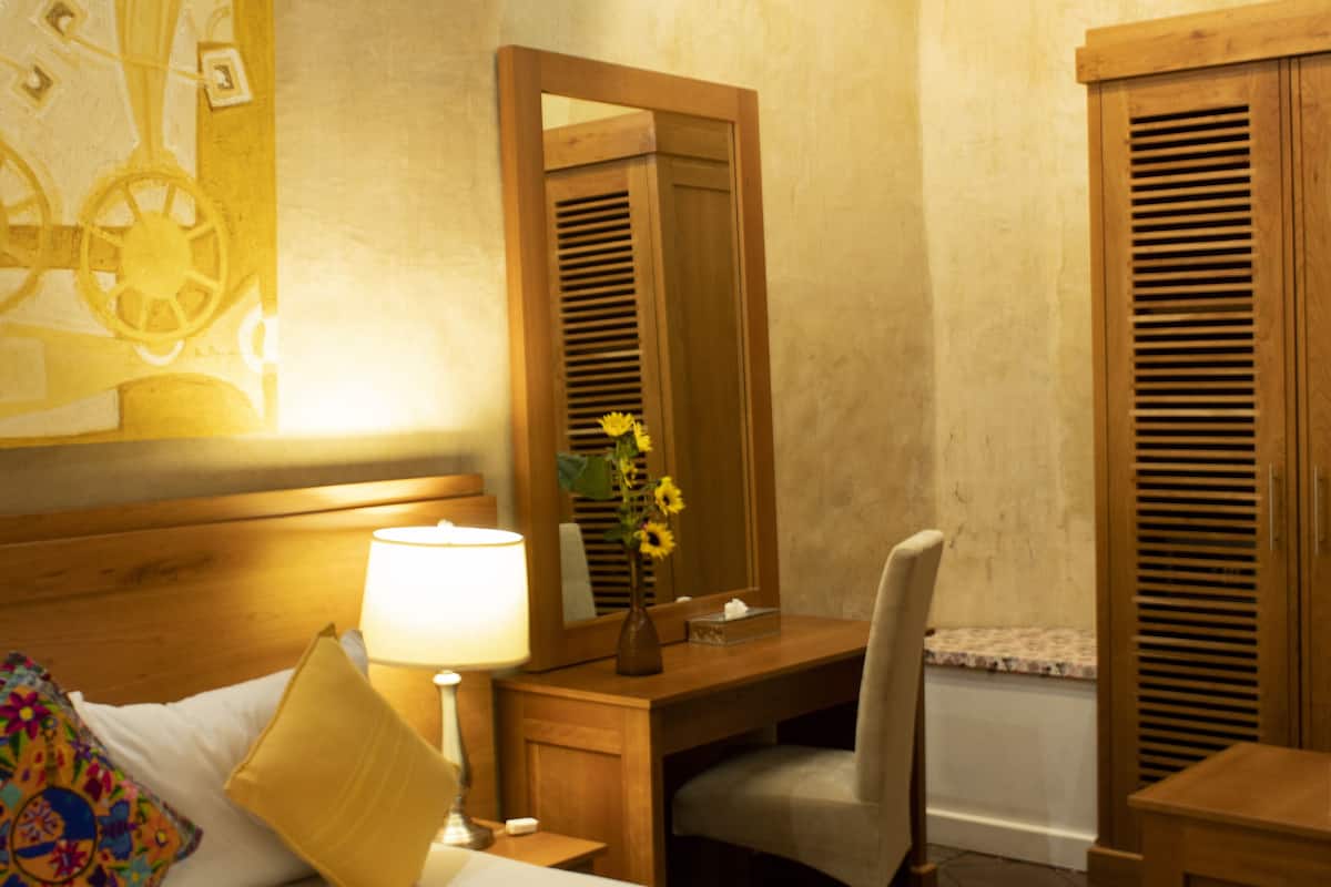 Habitación de hotel con cama, escritorio, silla, lámpara, espejo y un jarrón con flores.