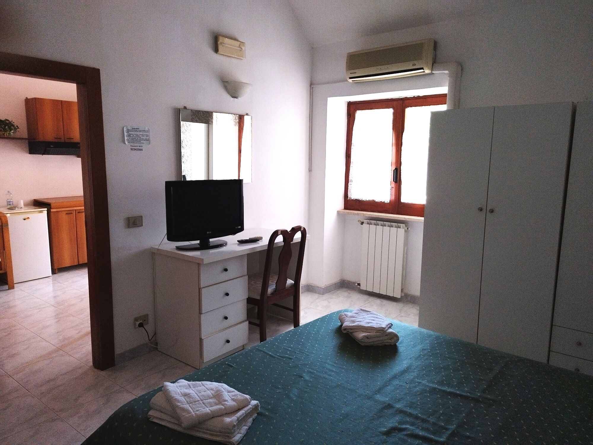 A small, tidy room with a desk, a chair, a bed with a green bedspread, a radiator, and an air conditioning unit.