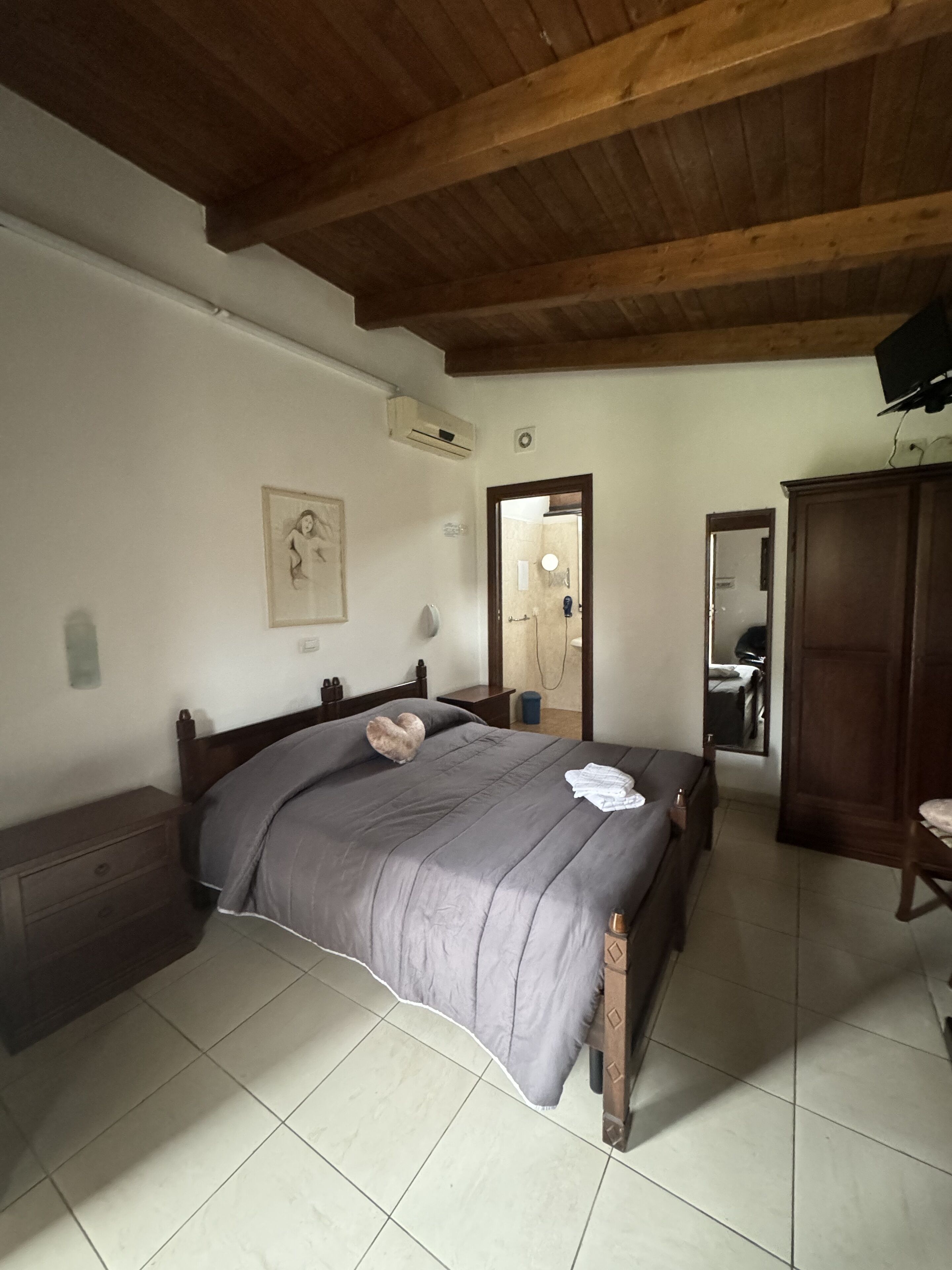 A bedroom with a wooden ceiling, a bed with a grey bedspread, a nightstand, a dresser, and a mirror.