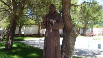 A statue of a person in a long robe standing on a pedestal in a park with trees and a stone wall in the background.
