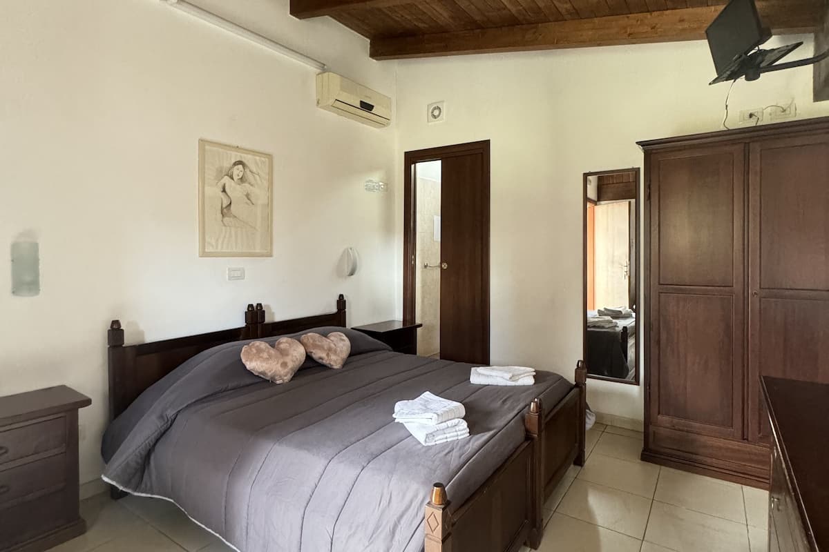 A bedroom with a wooden ceiling, a bed with a grey bedspread, a wooden wardrobe, and a wall-mounted air conditioning unit.