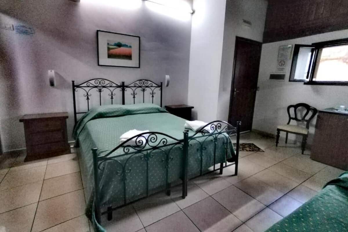 A bedroom with a metal bed frame, a green bedspread, a bedside table, a chair, and a window with a view of the outdoors.