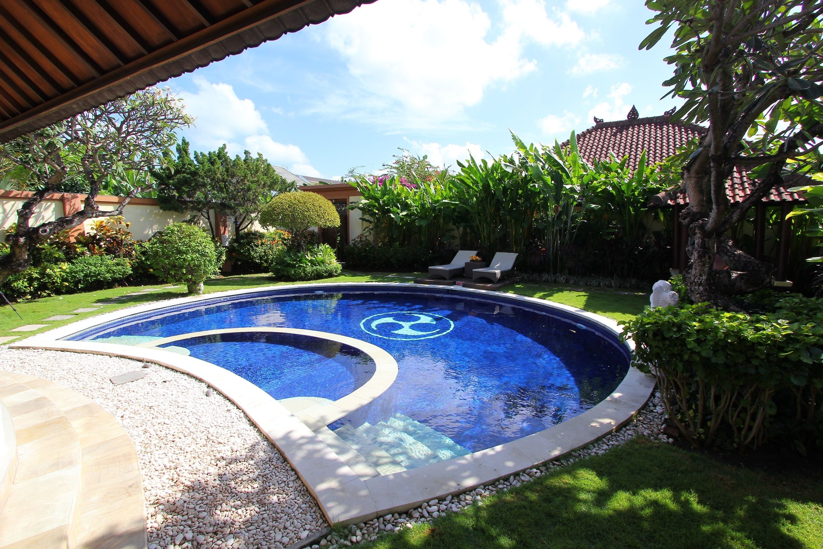 Une piscine de forme circulaire, entourée d’un jardin bien entretenu et surmontée d’une structure à toit recouvert de tuiles.