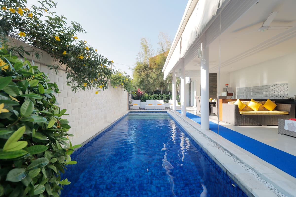 Una piscina con suelo de azulejos azules, rodeada por un muro blanco y abundante vegetación.