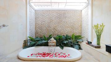 A bathroom with a stone wall, a bathtub filled with rose petals, and green plants.