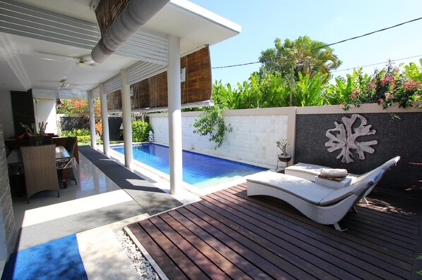 Un área junto a la piscina con una tumbona, una mesa y una planta en maceta.