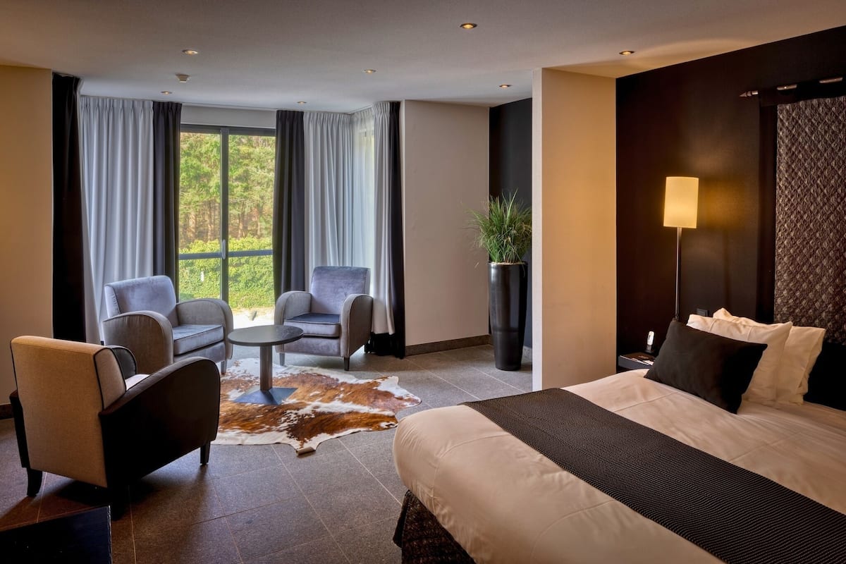 Een moderne hotelkamer met een groot bed, twee fauteuils, een tafeltje en uitzicht op het landschap door grote ramen.