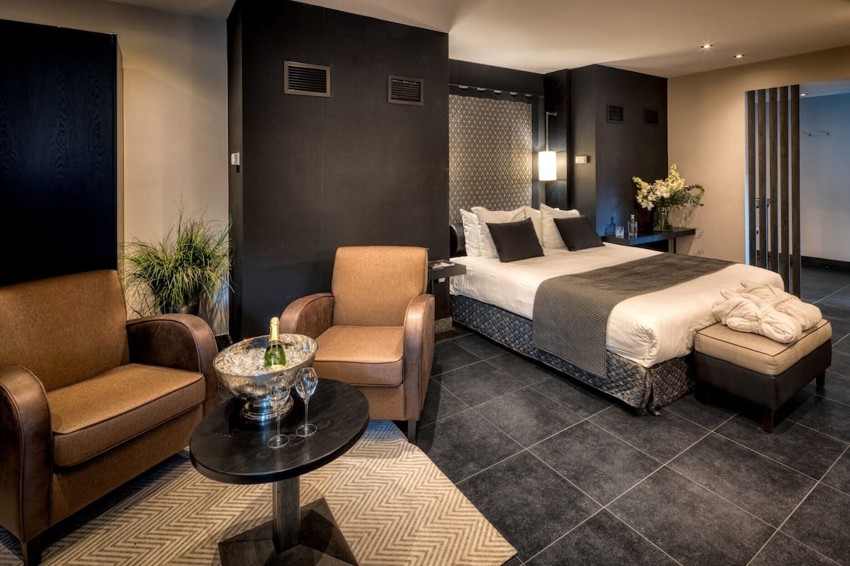 Een moderne hotelkamer met een bed, twee fauteuils, een tafeltje met een fles en glazen, en een plant in een pot.