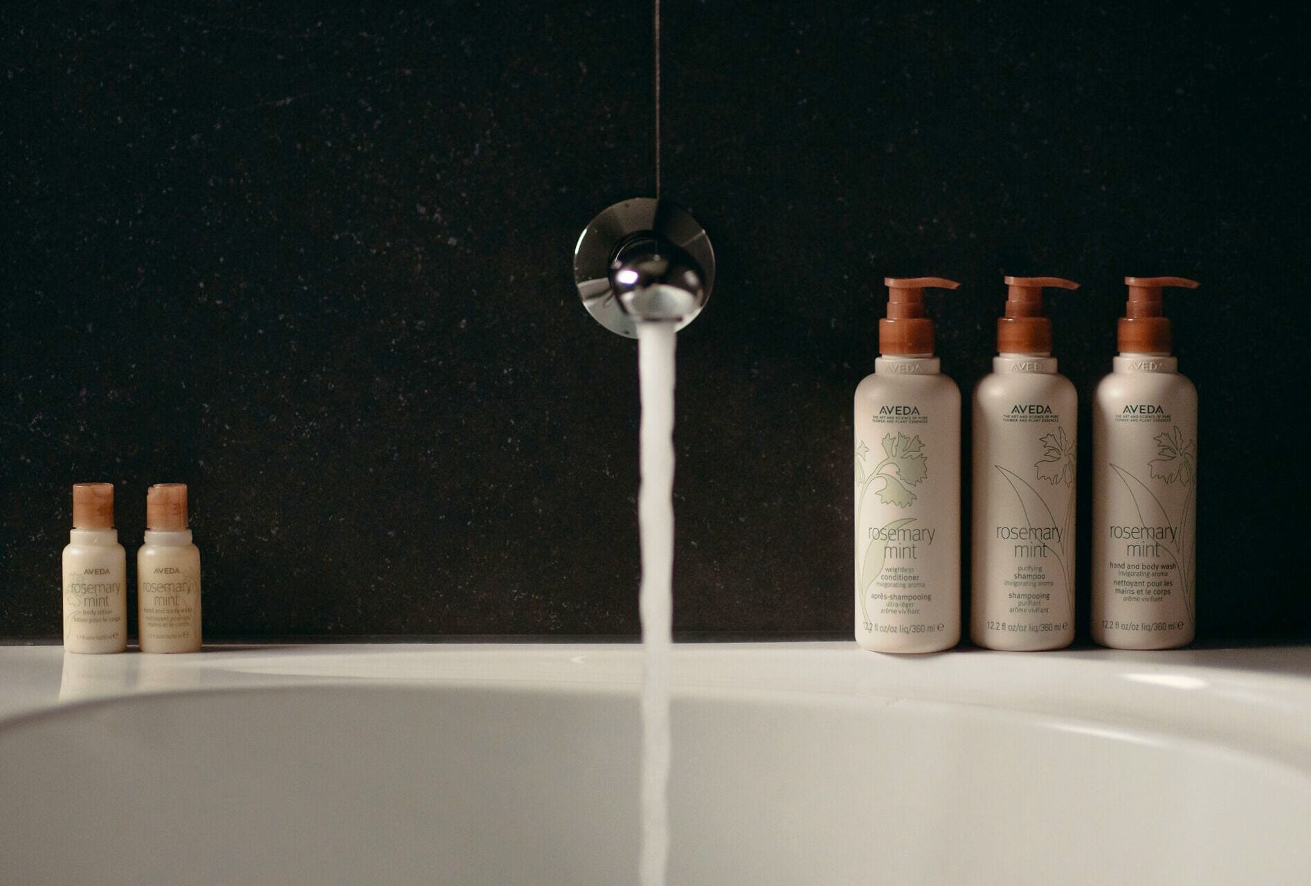 Un lavamanos de baño con cuatro botellas de champú y acondicionador de romero y menta Aveda, y un cabezal de ducha con agua que fluye hacia el lavamanos.