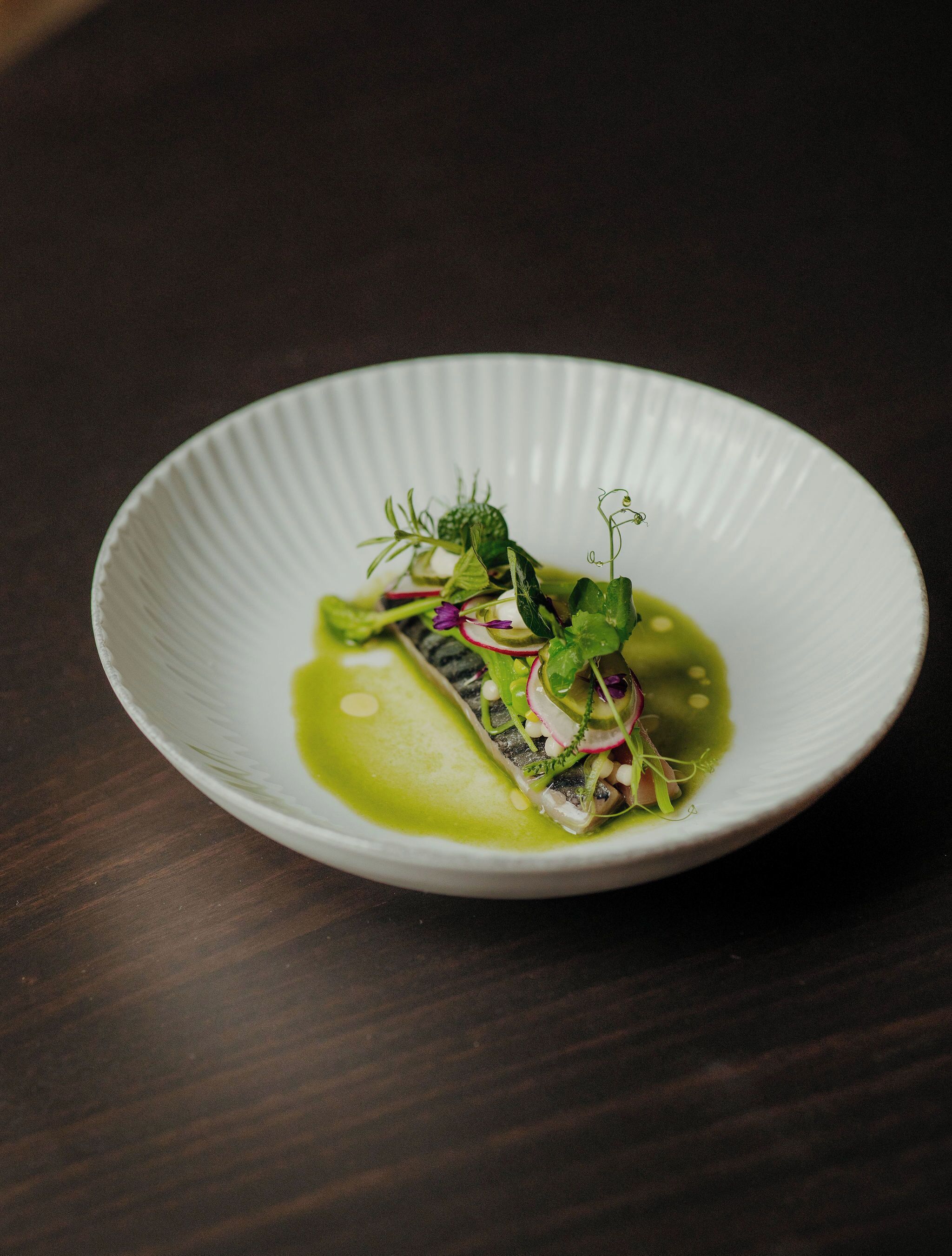Un plato blanco con salsa verde, pescado y verduras sobre una mesa de madera oscura.