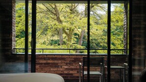Habitación con un amplio ventanal que da a un exuberante jardín verde. Se ve una cama y dos sillas.