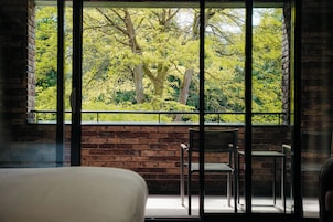 Habitación con un amplio ventanal que da a un exuberante jardín verde. Se ve una cama y dos sillas.