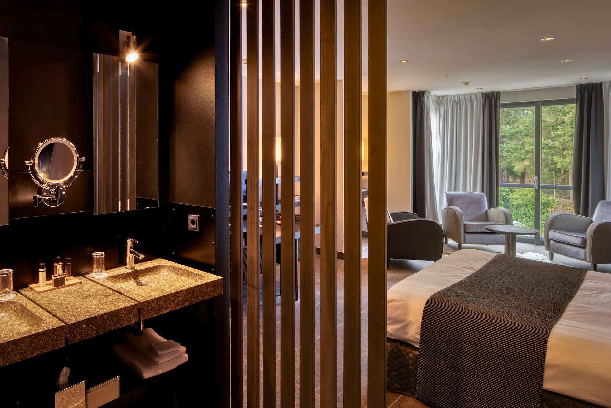 A hotel room with a double bed, a sitting area with two chairs, a small table, a bathroom with a double sink, and a large window with curtains.