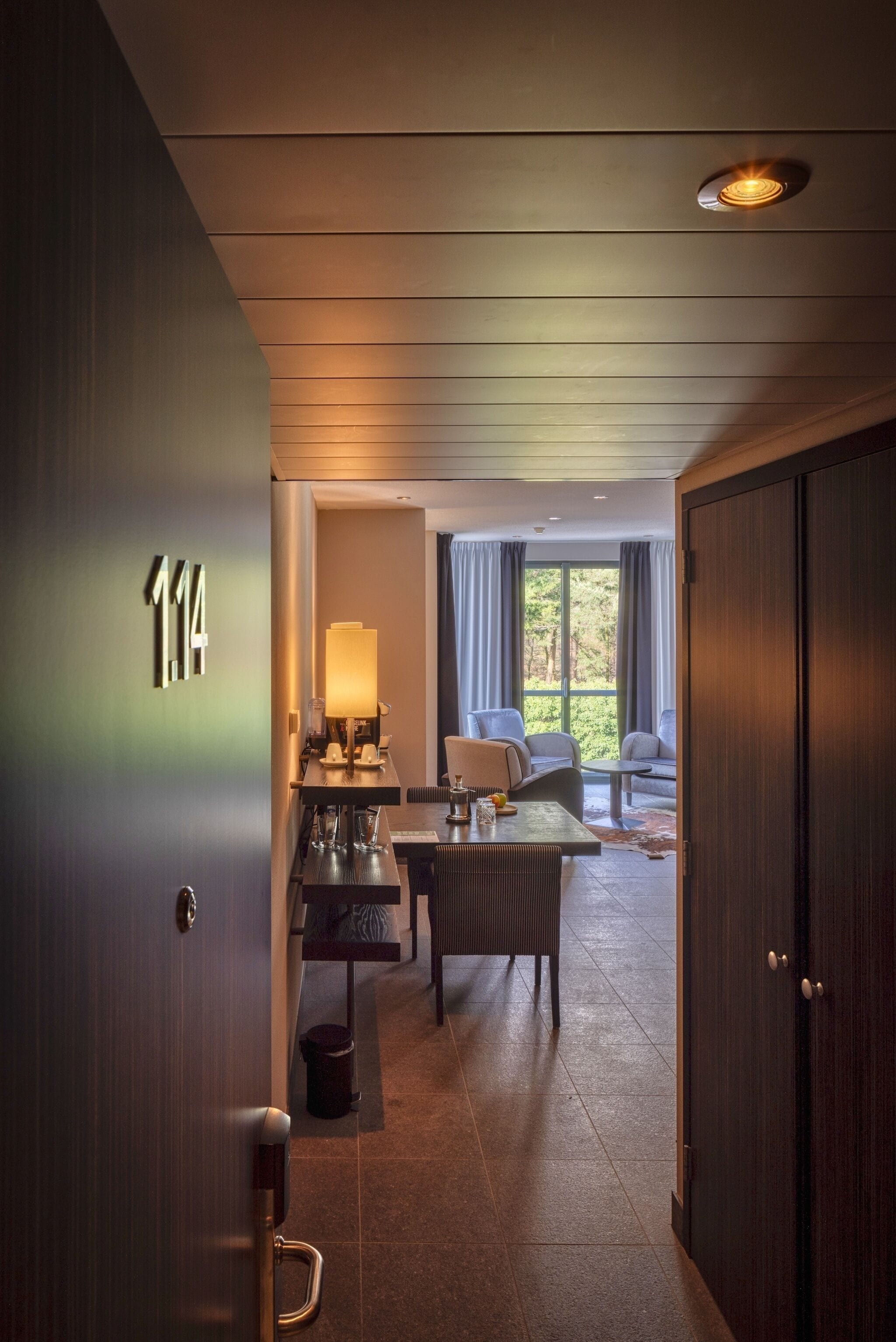 A hotel room with a wooden door marked "114", a desk with a lamp, a sofa, and a view of the outdoors through large windows.