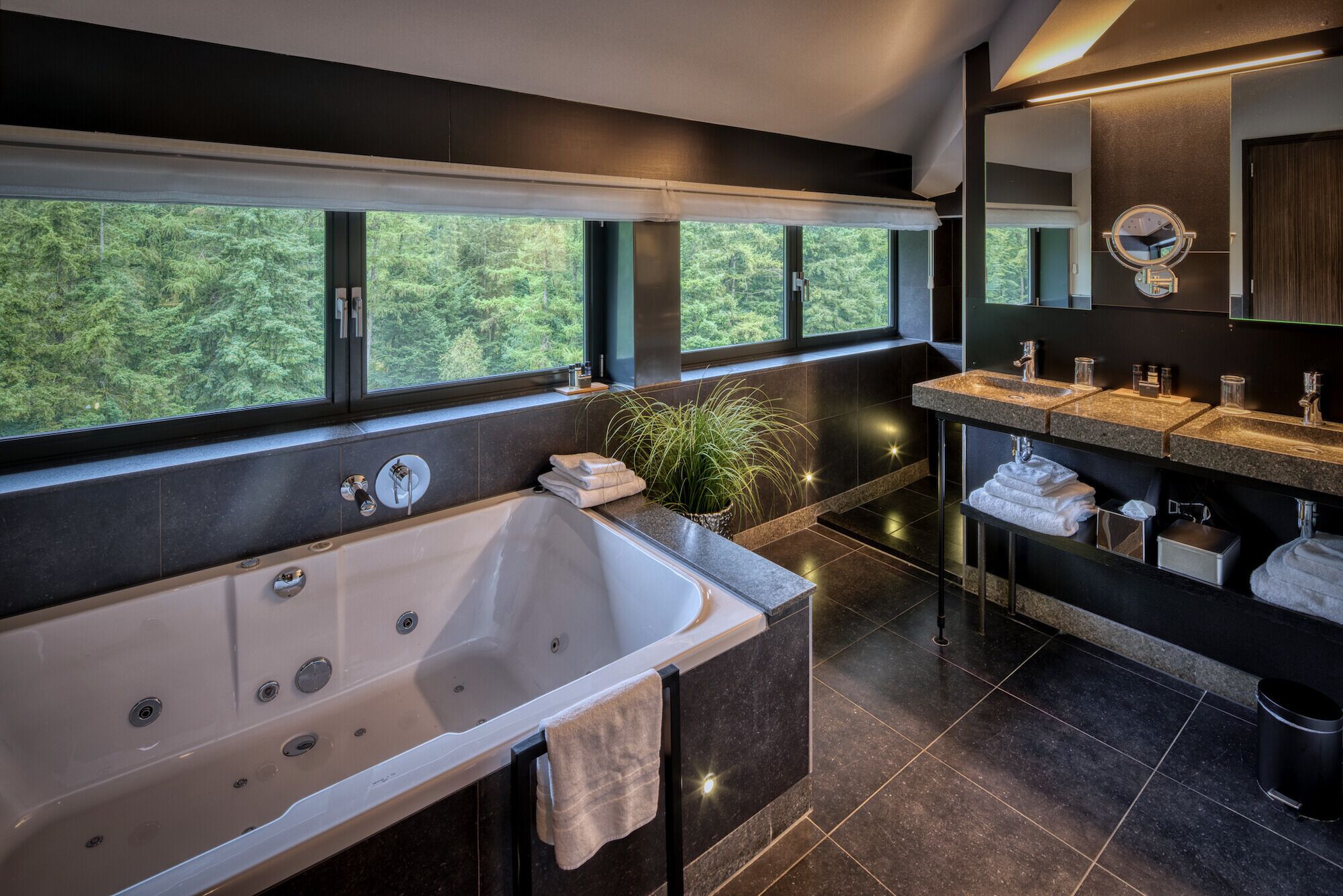 Un baño moderno con una bañera grande, un tocador con lavamanos y un espejo. Hay ventanas que ofrecen vistas al bosque.