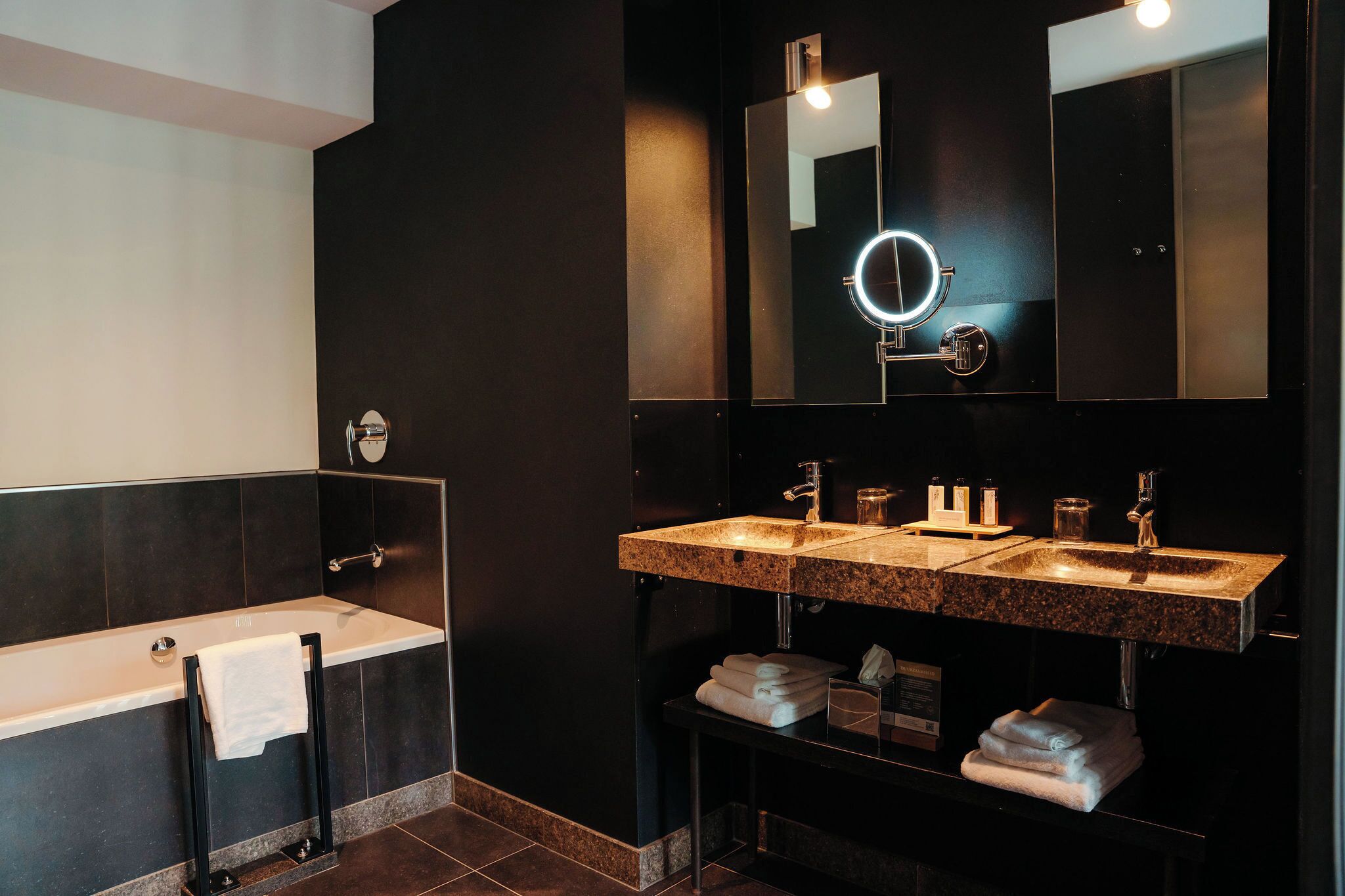 A modern bathroom with a bathtub, two sinks, a vanity, and a large mirror with a light.
