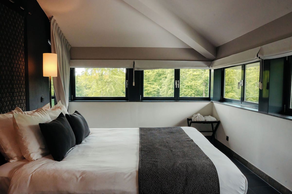 Een hotelkamer met een groot bed, een nachtkastje, een lamp en uitzicht op bomen door de ramen.