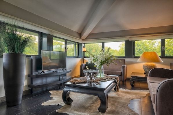 A modern living room with a large TV, comfortable seating, a coffee table, and a view of greenery through large windows. - Hotel de Echoput (Hoog Soeren)