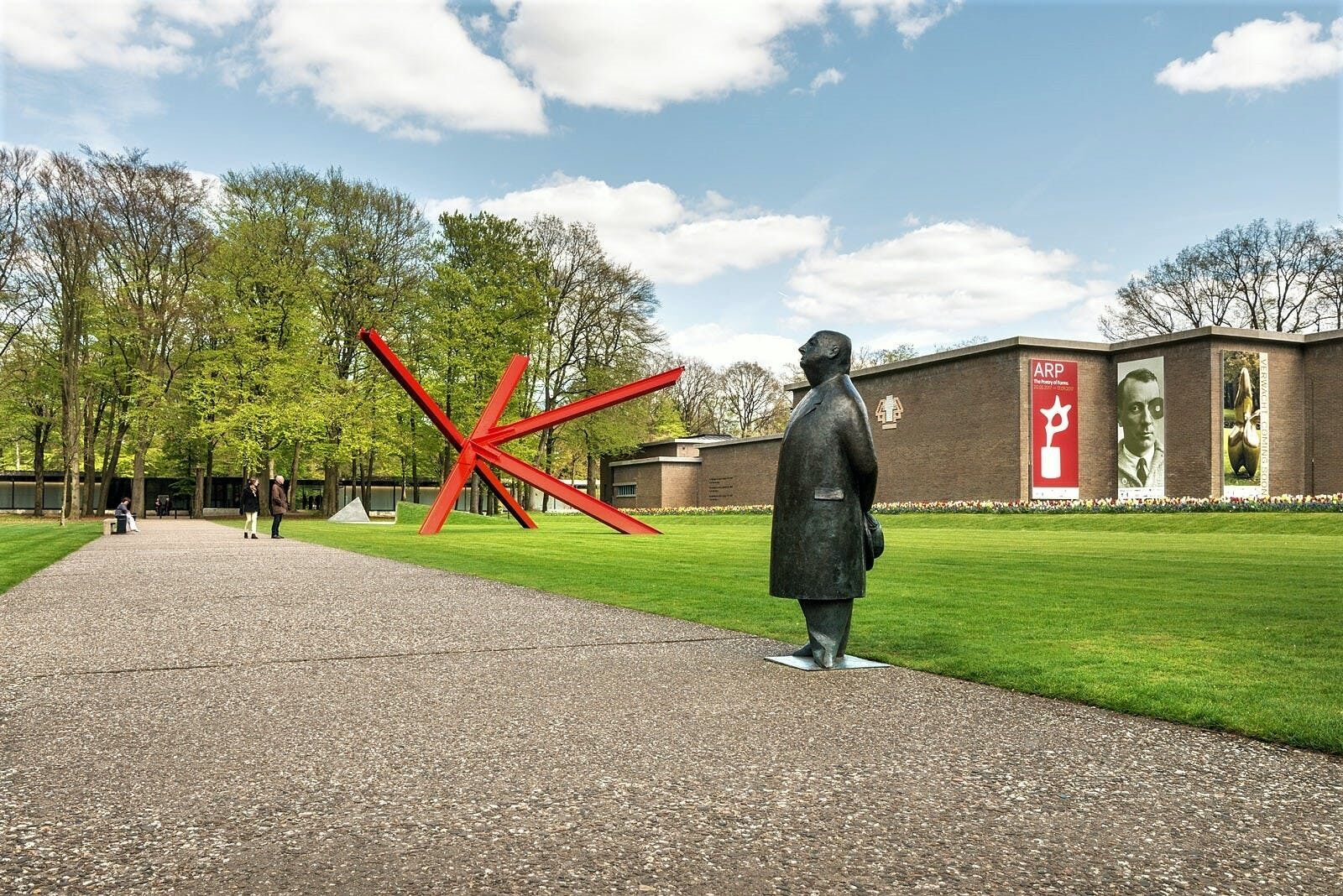 A statue of a man in a suit standing on a paved path with a red sculpture in the background.