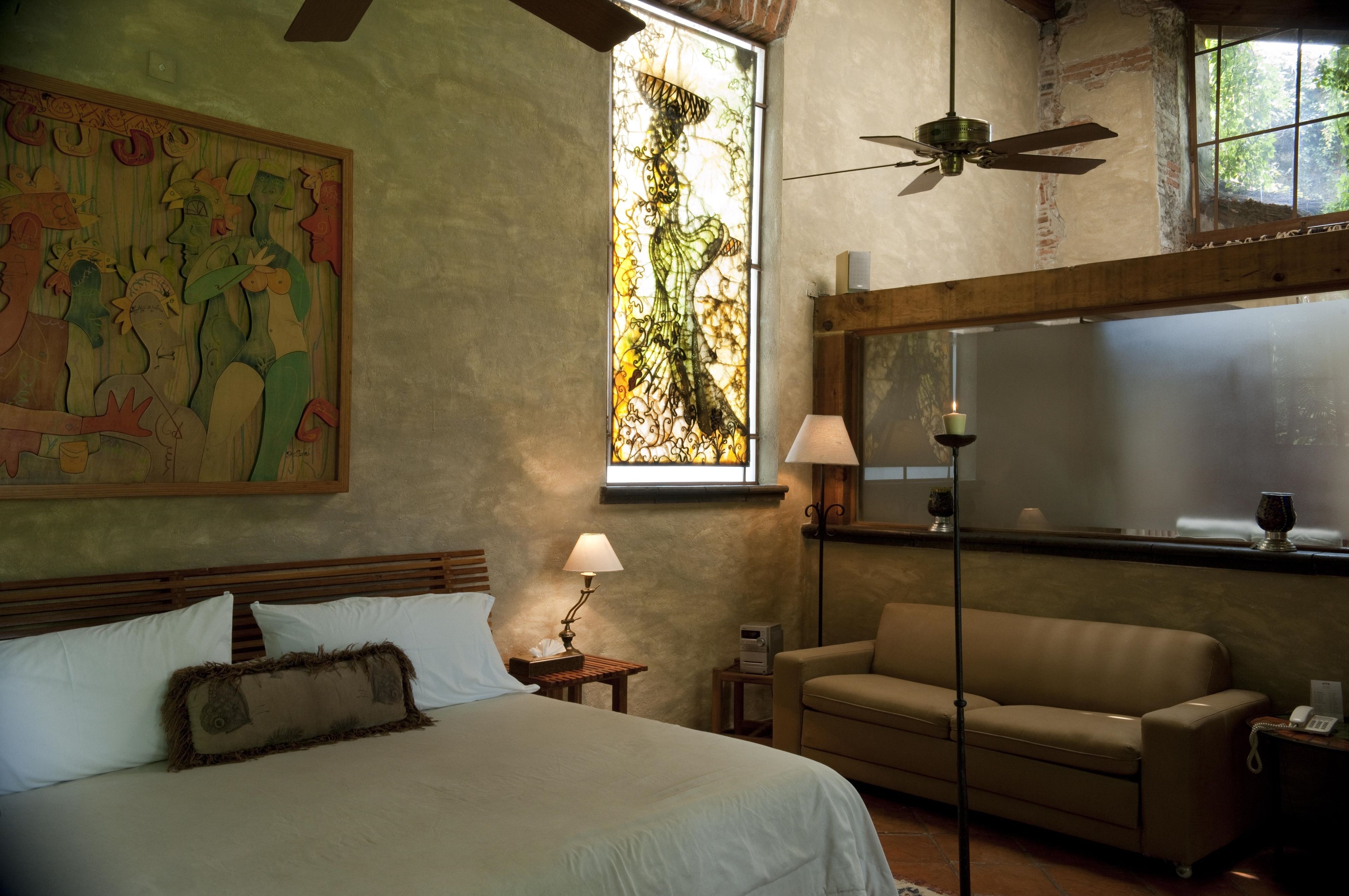 A hotel room with a large bed, a sofa, a window with a view of trees, a ceiling fan, and a painting on the wall.