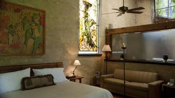 A hotel room with a large bed, a sofa, a window with a view of trees, a ceiling fan, and a painting on the wall.
