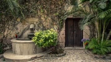 A stone fountain surrounded by lush greenery and a stone pathway leading to a dark wooden door.