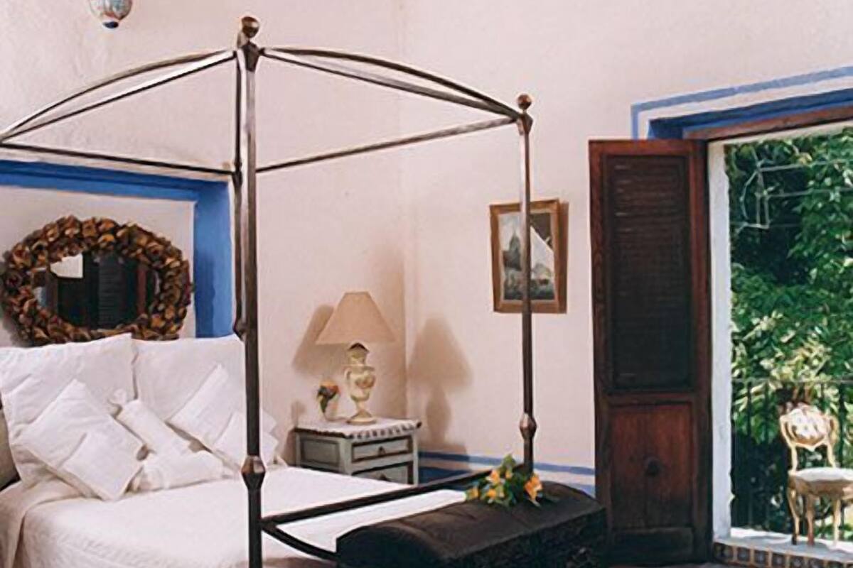 Una cama con dosel, una mesita de noche con lámpara, un cuadro en la pared y una puerta que da a un jardín.