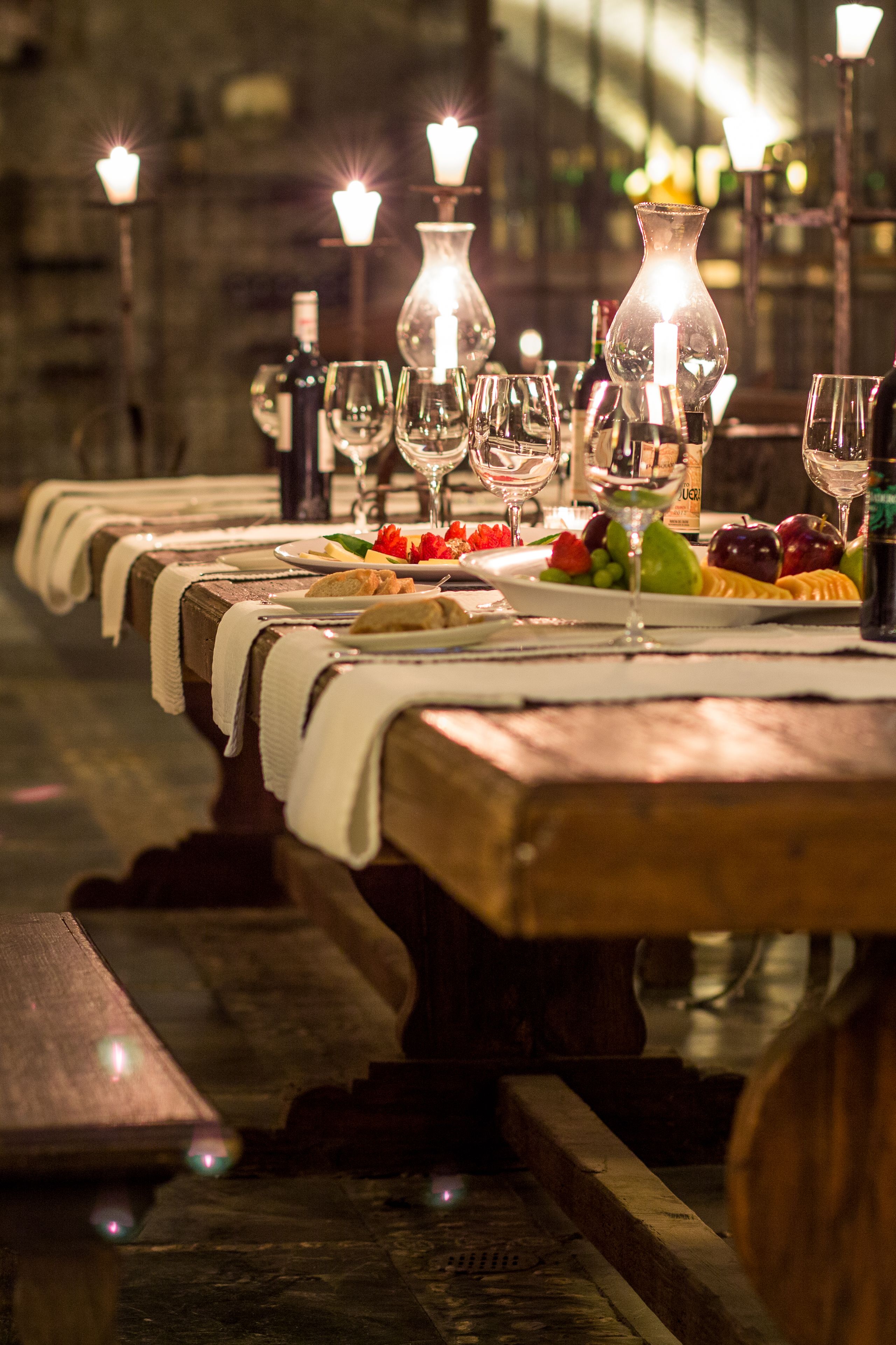 Una larga mesa de madera dispuesta para comer, con velas, copas de vino y una variedad de platos.