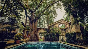 Una piscina rodeada de abundante vegetación y una estructura de piedra con un ventanal circular.
