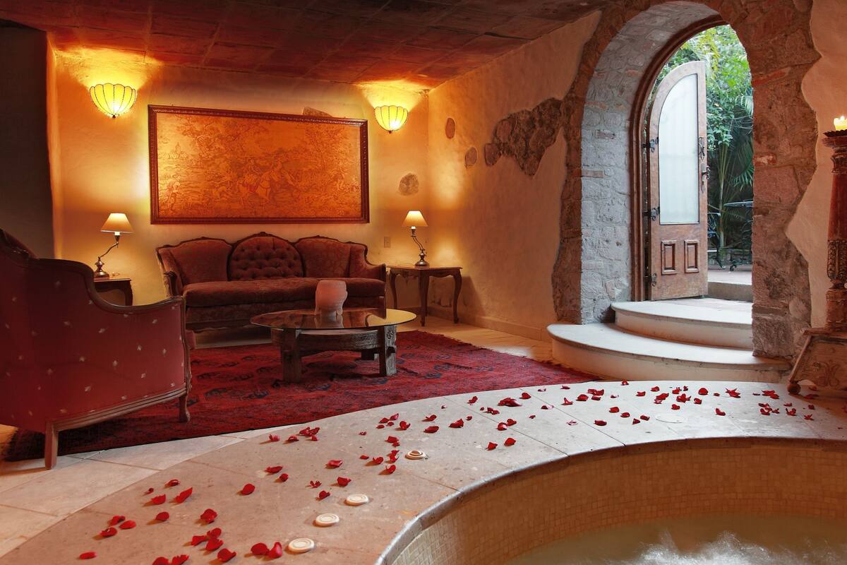 Una habitación con una bañera de piedra llena de agua, una alfombra roja con pétalos de rosa, un sofá rojo y un cuadro en la pared.