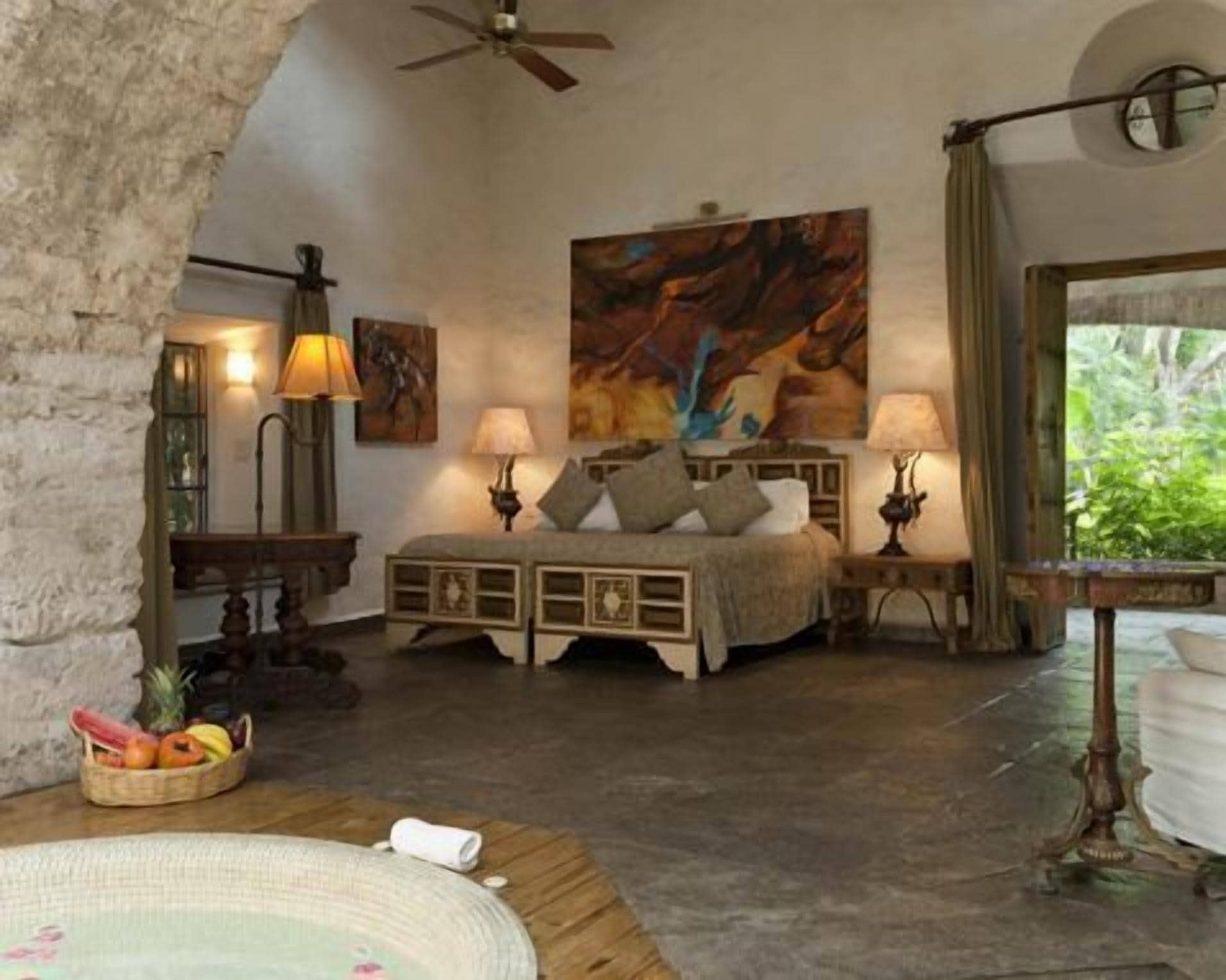 A cozy bedroom with a bed, two bedside tables with lamps, a large painting on the wall, and a view of greenery outside.