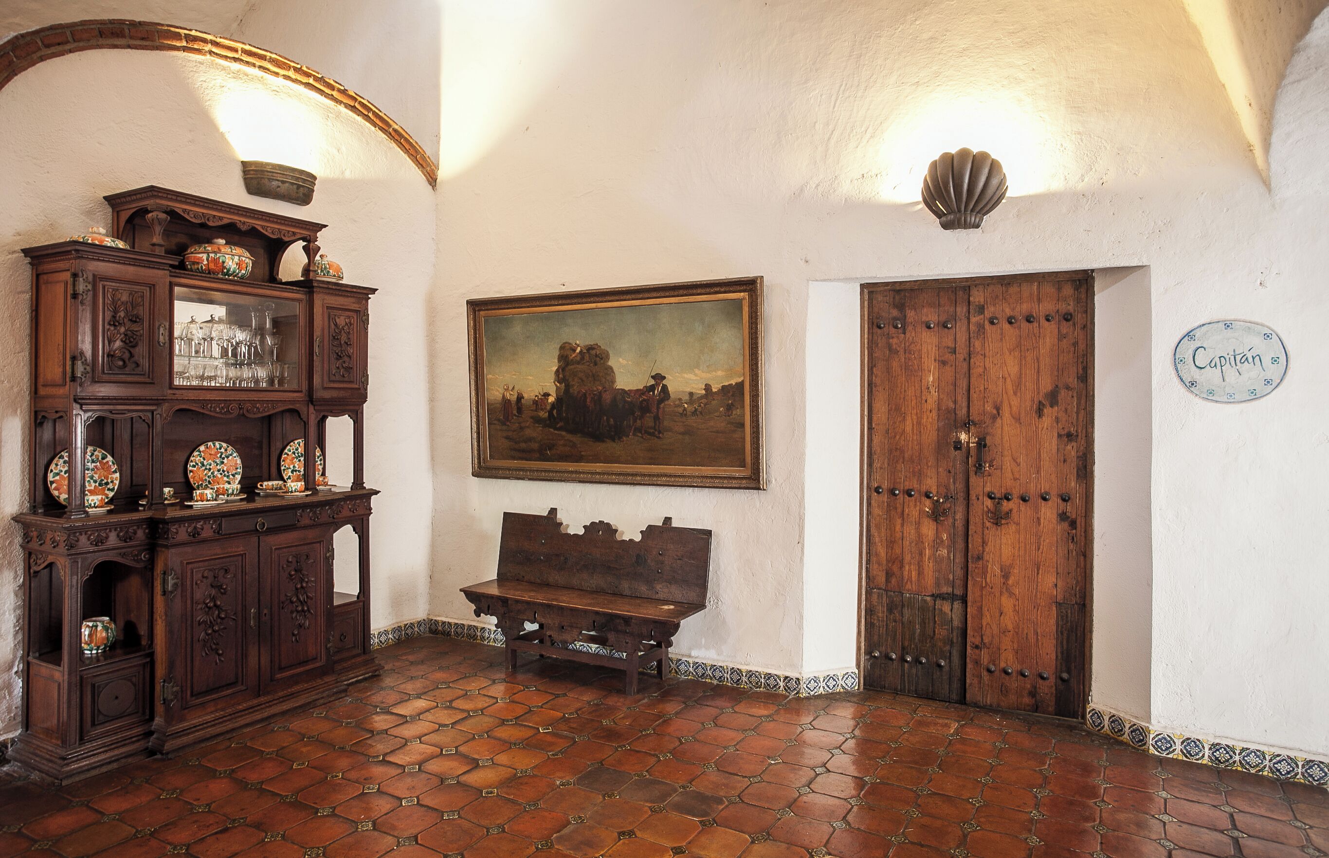 A room with a wooden cabinet, a bench, a painting, and a door labeled "Capitan".