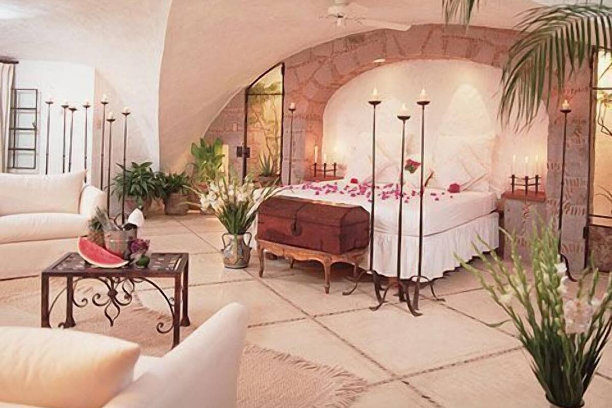 Un dormitorio con un arco de piedra, una cama con pétalos de rosa, una mesita de noche, un aparador, un sofá, una mesa de centro y una planta en maceta.