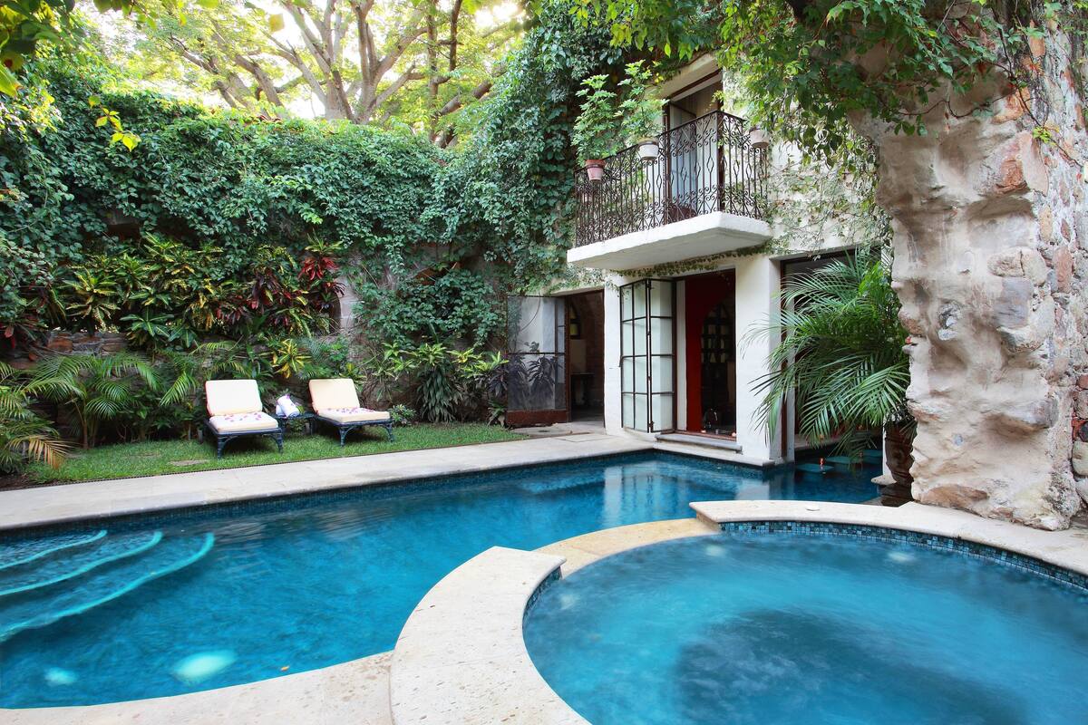 Una piscina de borde curvo, rodeada de abundante vegetación y un muro de piedra.