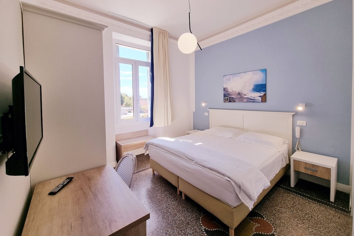 Un dormitorio con cama, mesita de noche, televisor, ventana con cortinas y un cuadro en la pared.