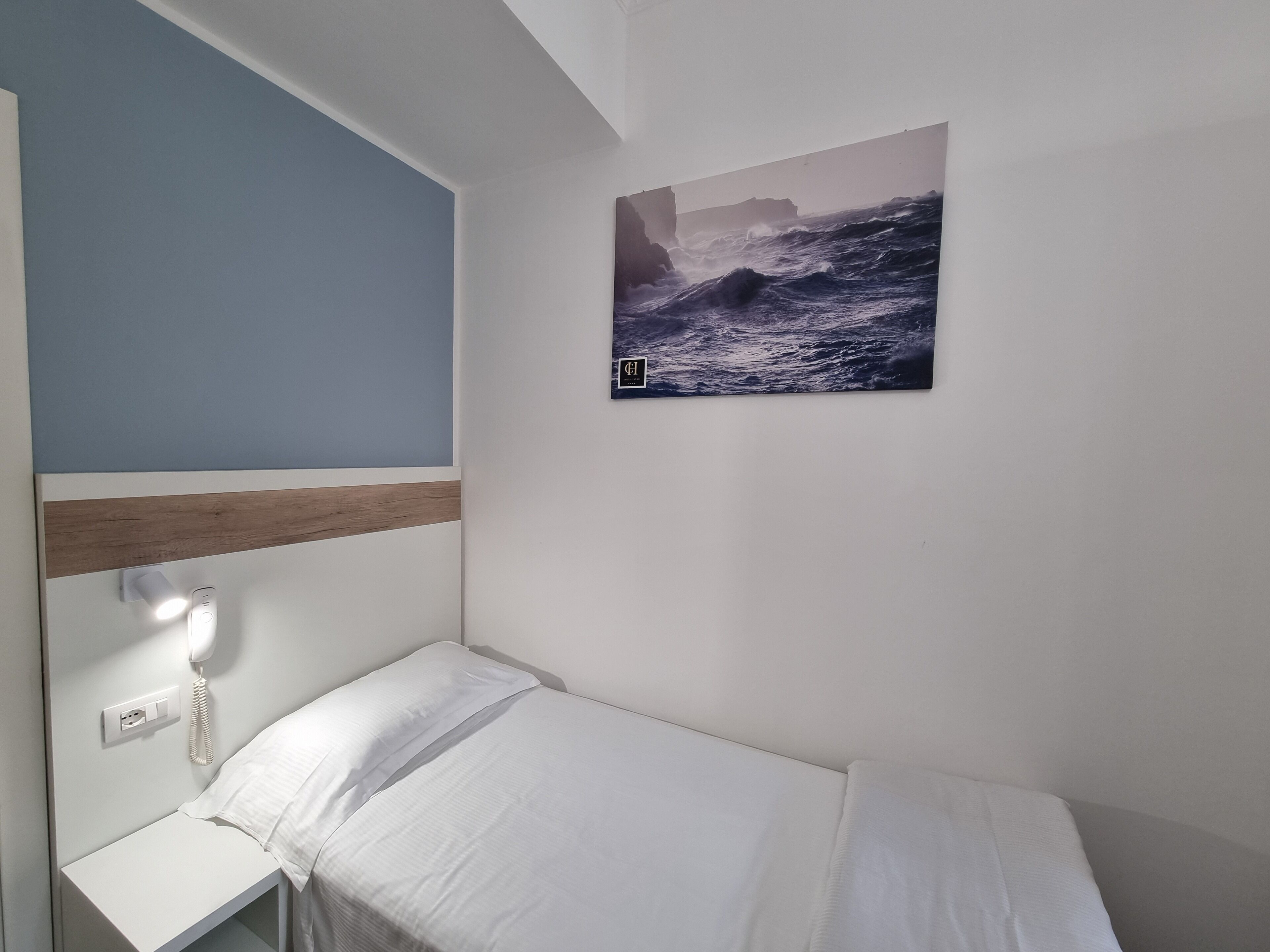 A small, neatly made bed in a room with a single bedside table, a mounted telephone, and a framed picture of a seascape on the wall.