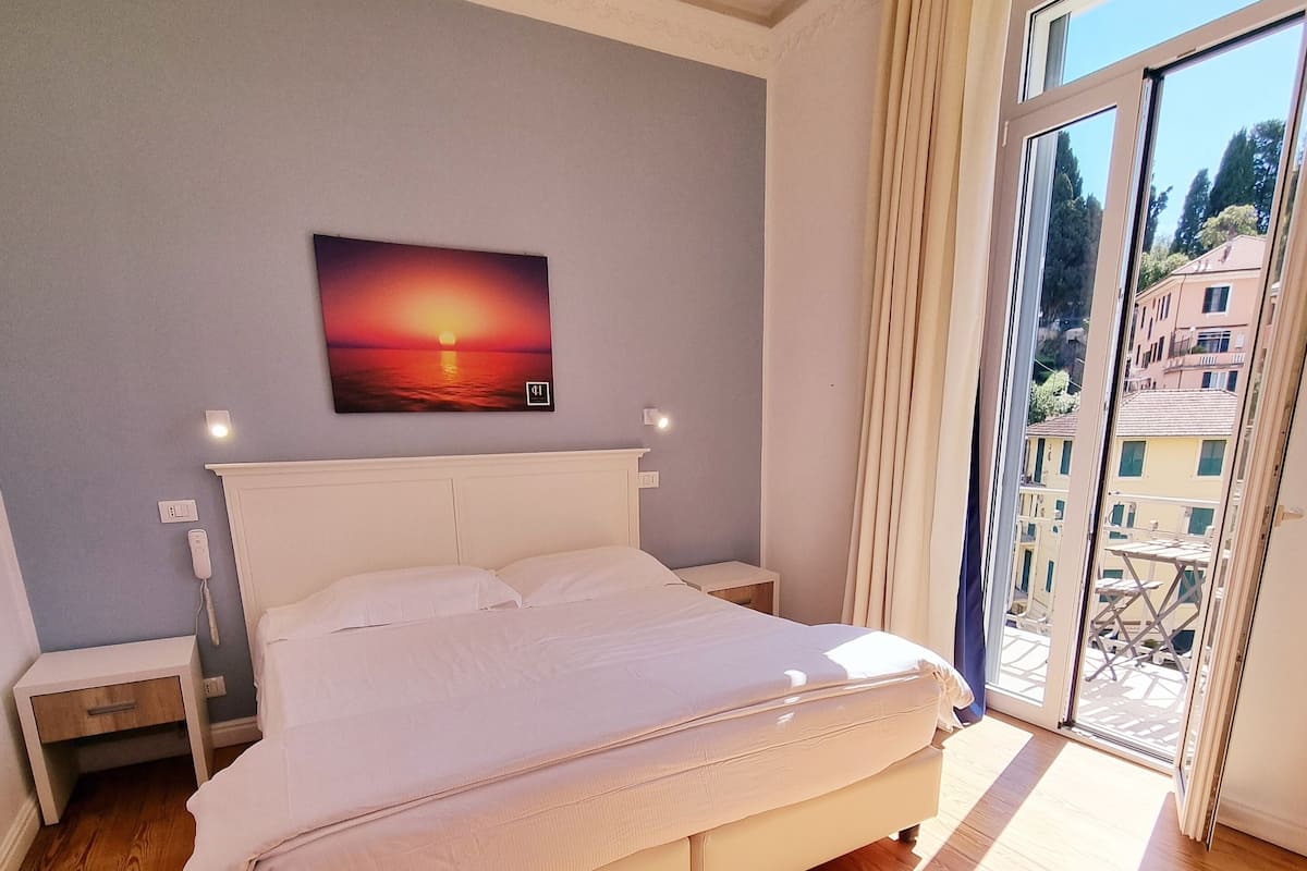 Un dormitorio con una cama grande, suelo de madera, un cuadro de un atardecer y vistas de edificios a través de una puerta de vidrio.