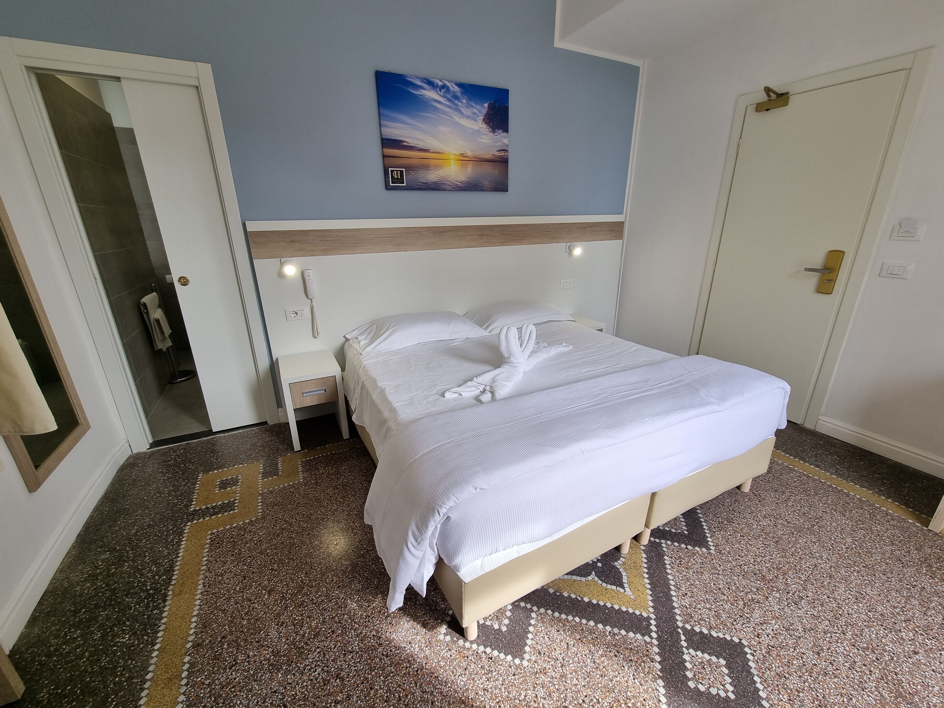 A hotel room with a bed, a nightstand, a wall-mounted phone, a door, and a patterned floor.