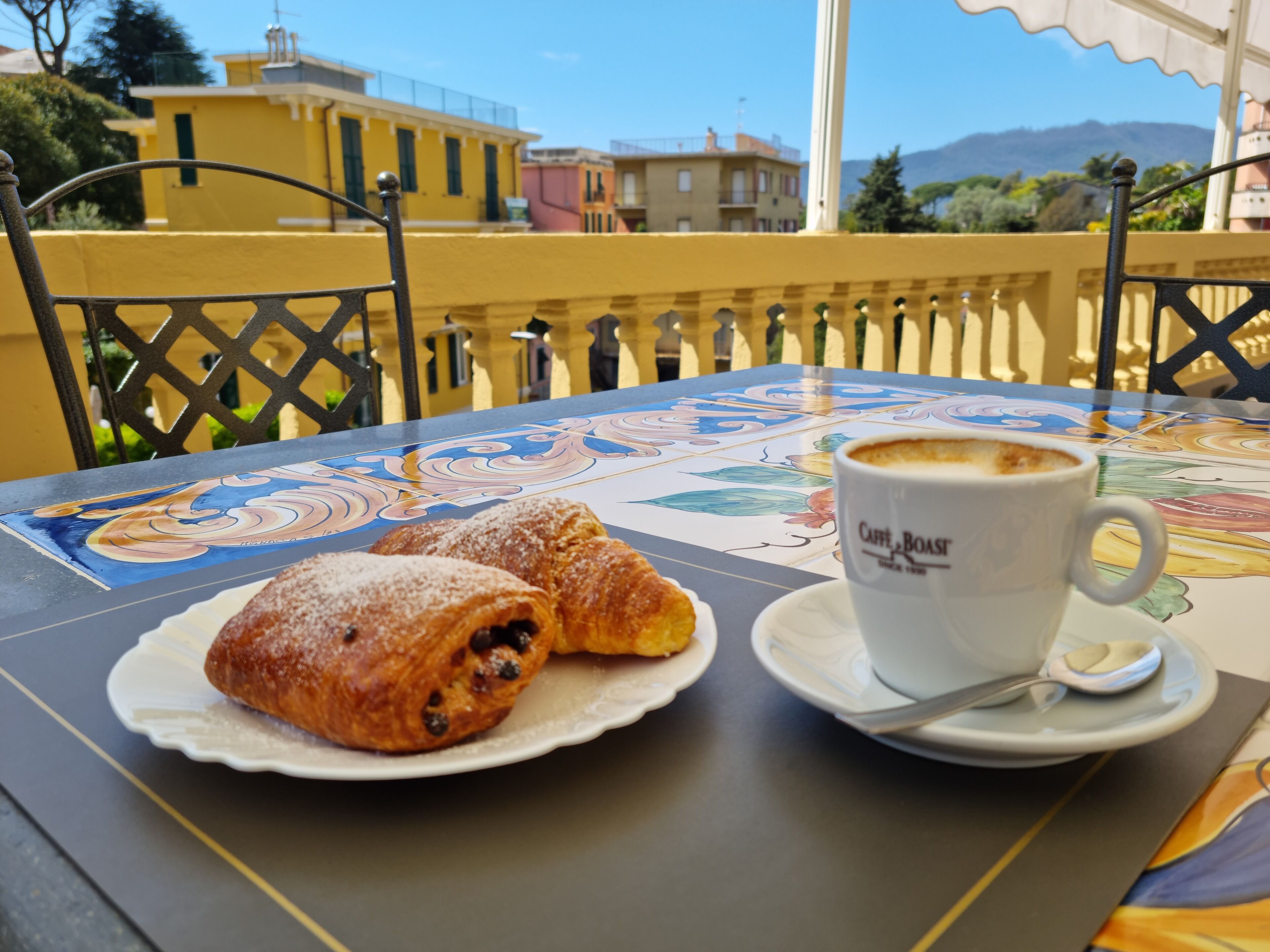 Et bord med et bagværk og en kop kaffe foran bygninger og et bjerg.