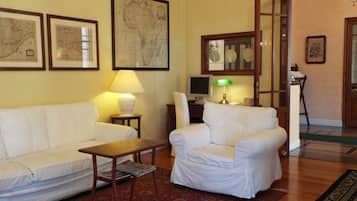 Un salón con un sofá blanco, un sillón blanco, una mesita y una lámpara. Hay mapas enmarcados en la pared y un ordenador sobre un escritorio.