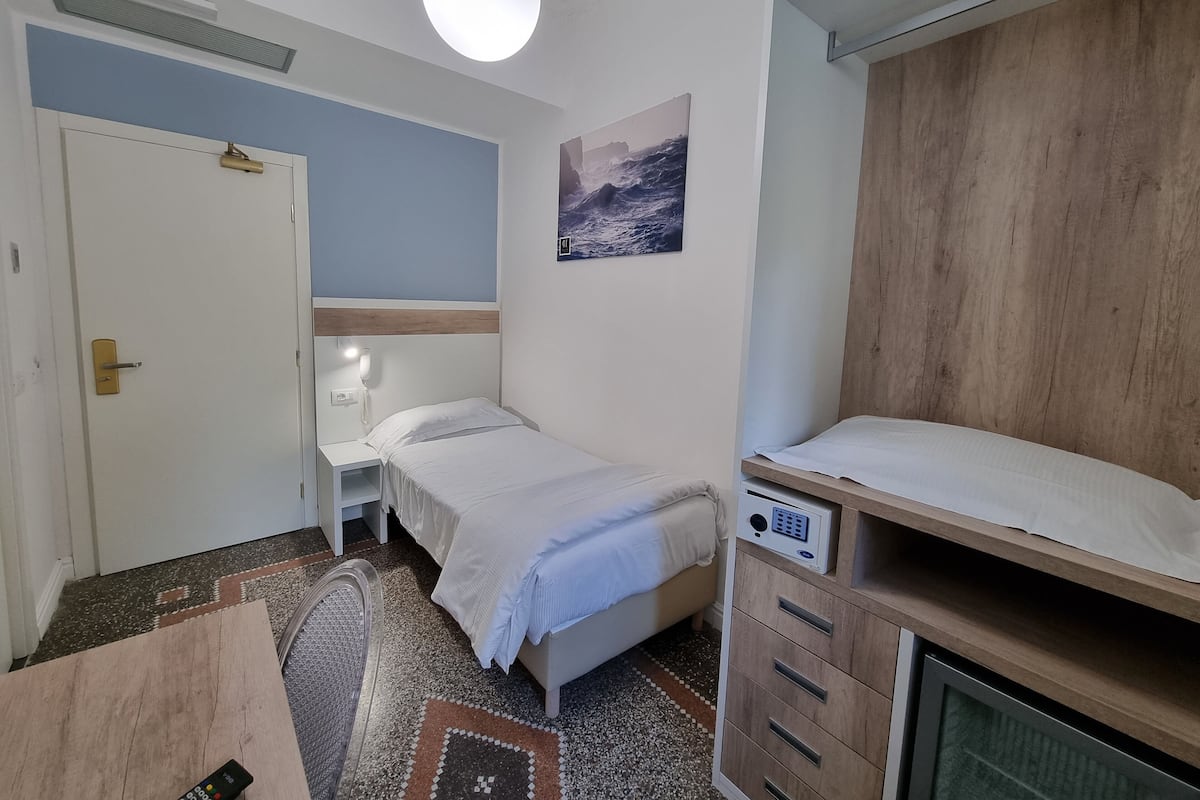 Un dormitorio con cama, mesita de noche, escritorio, silla, armario, teléfono de pared y un cuadro.