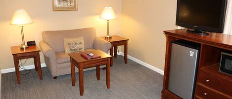 A hotel room with a sofa, two bedside tables with lamps, a coffee table, a television on a wooden cabinet, and a refrigerator.