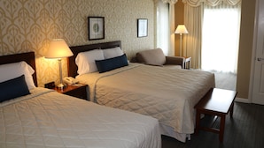 A hotel room with two beds, a bedside table with a lamp, a chair, and a small table.