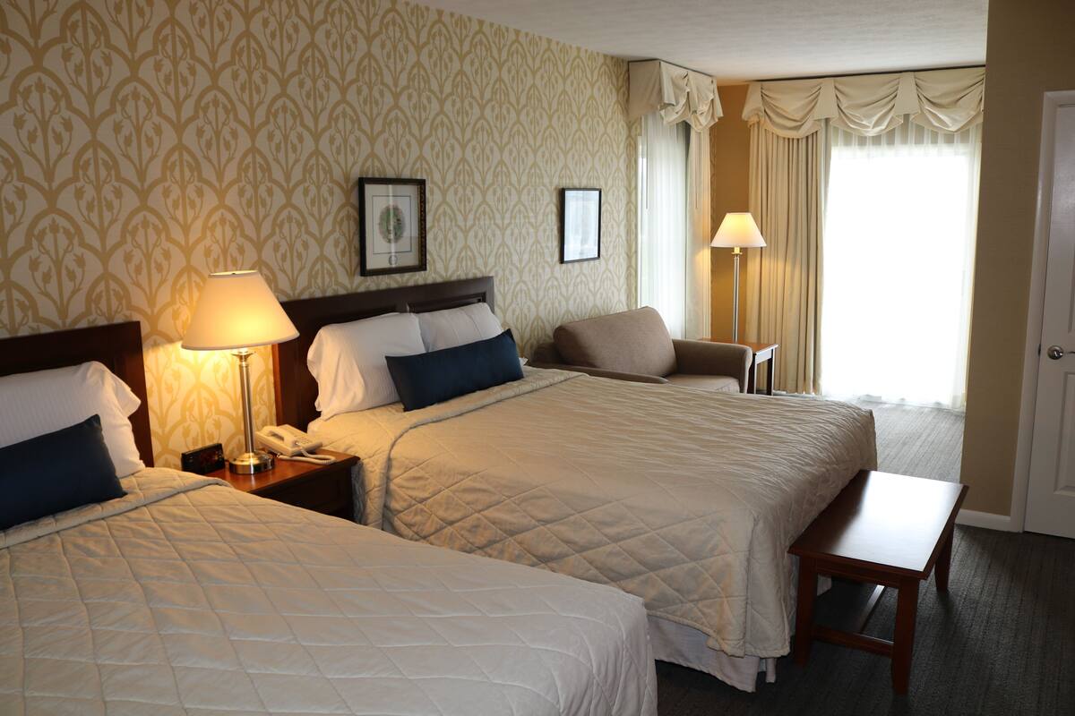 A hotel room with two beds, a bedside table with a lamp, a chair, and a small table.