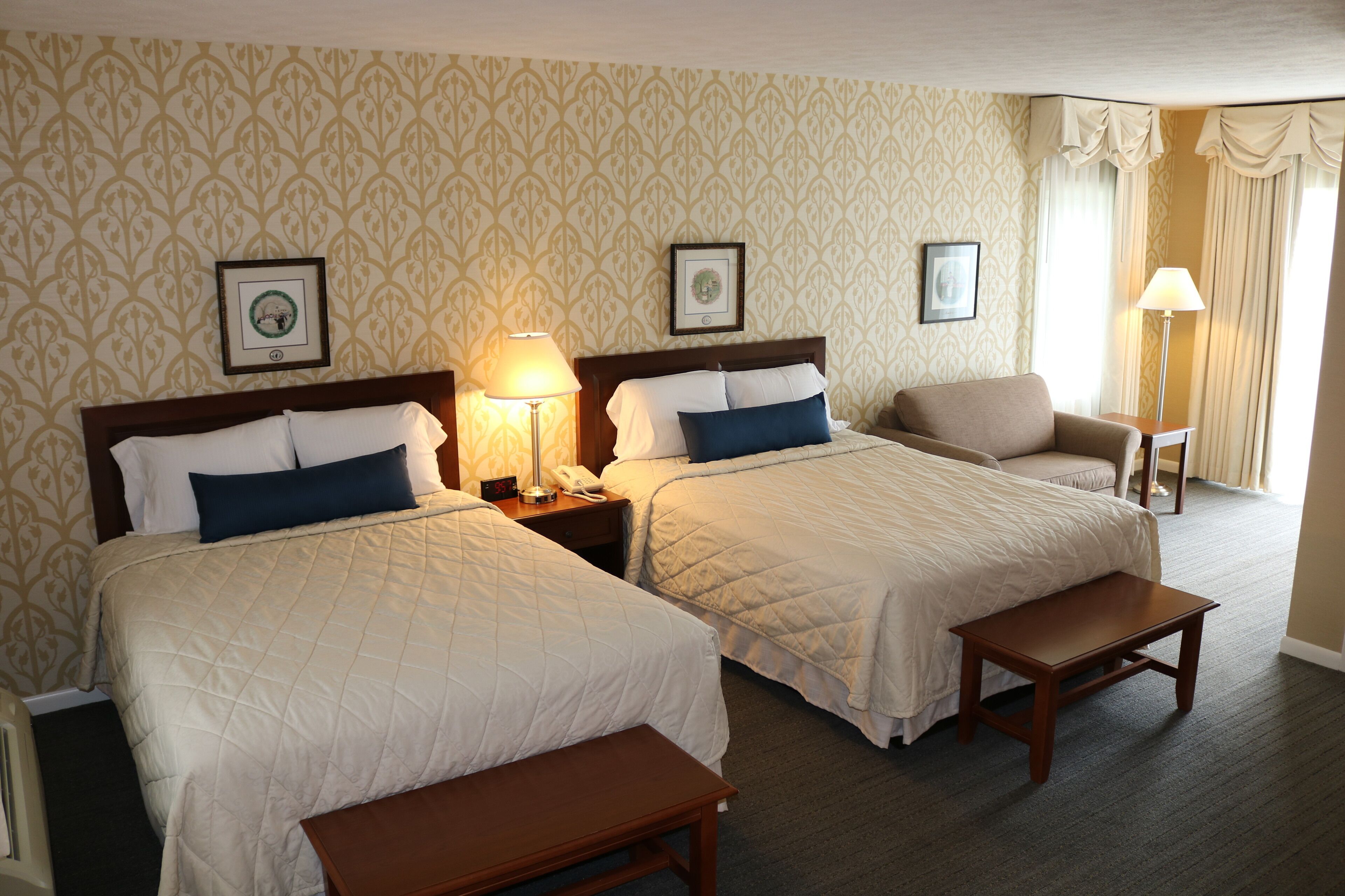Habitación de hotel con dos camas tamaño queen, un banco de madera, una mesita de noche con lámpara y una silla.