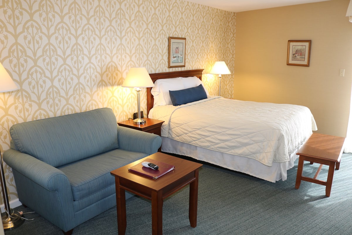 A hotel room with a bed, a sofa, a bedside table with a lamp, and a small wooden bench.